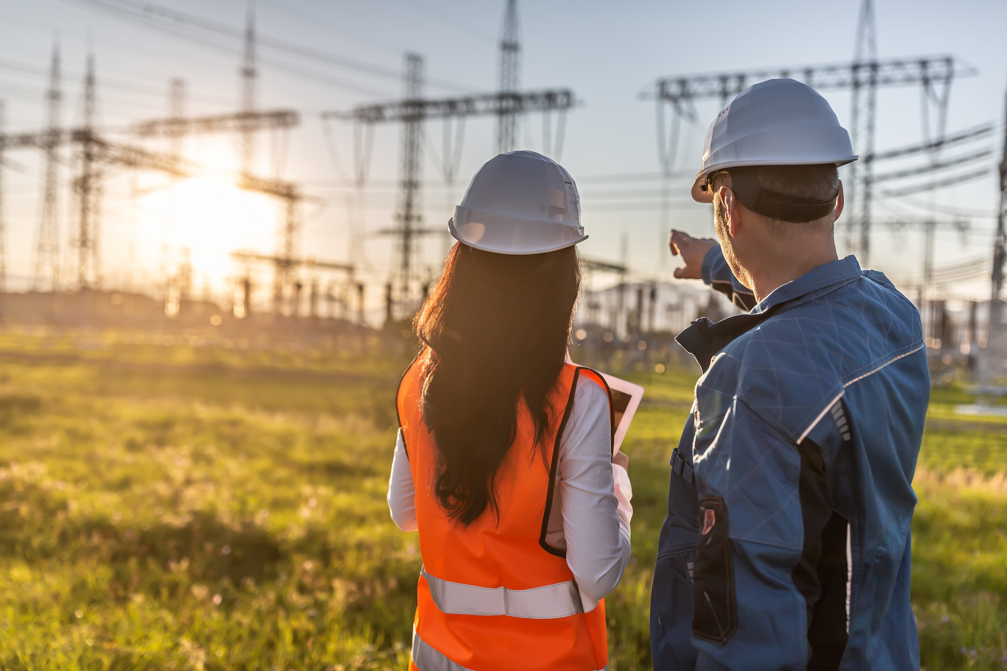 Blue collar worker points towards electricity poles with sun setting over it, with a female enginners on his side