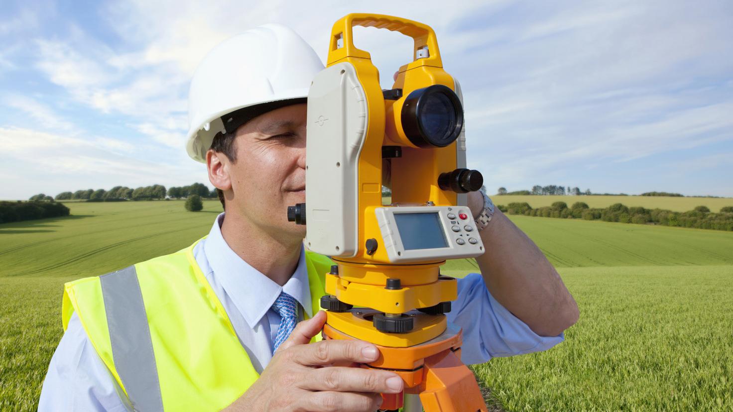 A man in a construction hat and vest stares into a surveying instrument