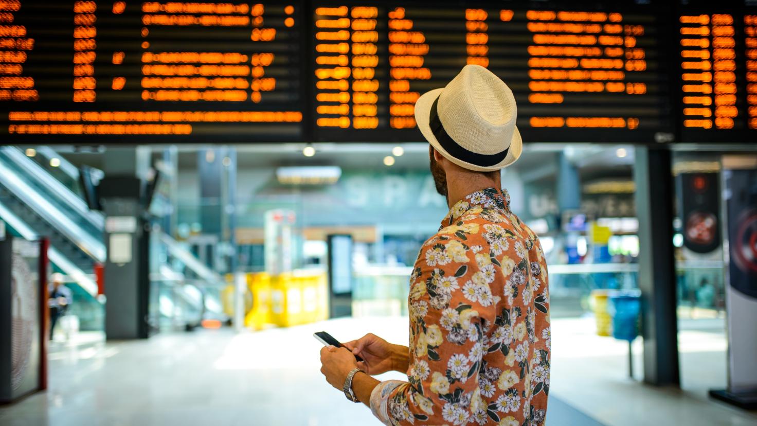 Ein Mann mit Hut schaut auf eine Abflugtafel in einem Bahnhof
