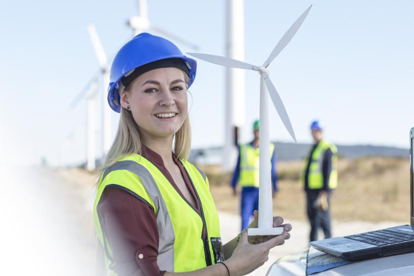 Eine Frau in einer fluoreszierenden Weste und einem Schutzhelm hält ein Modell einer Windkraftanlage