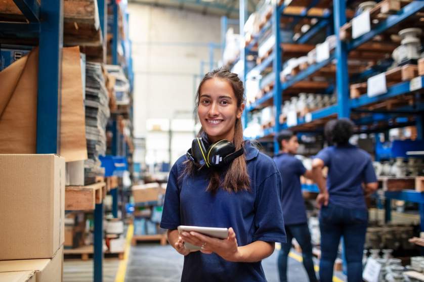 Portrait of a female factory employee with a digital tablet looking at camera and smiling. Woman warehouse worker with people in background.