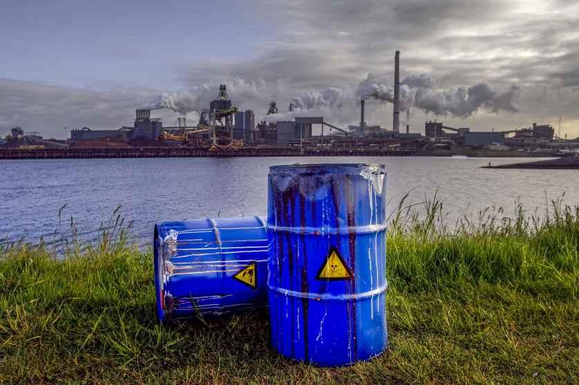 Empty Blue Chemical Waste Drums Lying on an Abandoned Bank with a view on Smoking Exhaust Pipes of a Heavy Industrial Factory