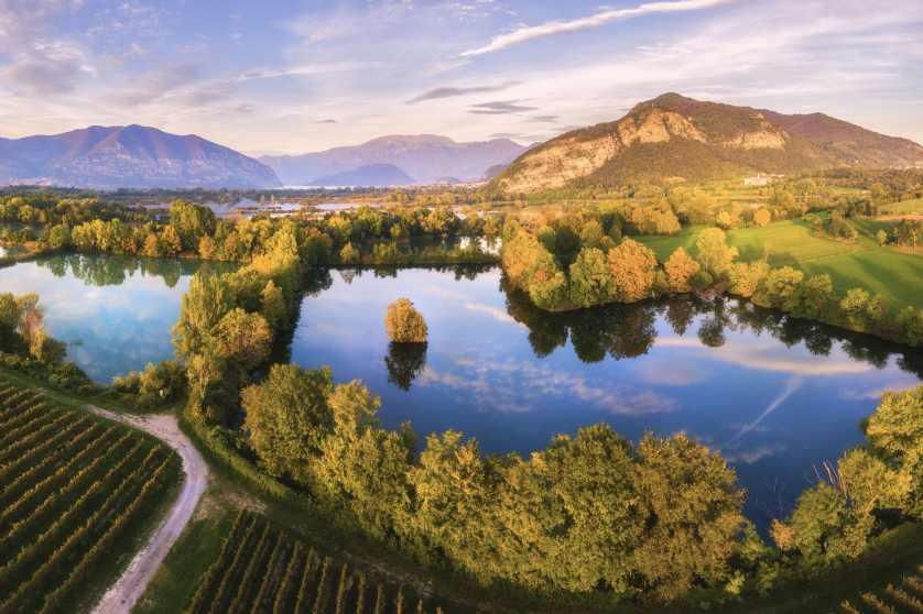 Letecké panorama malebného říčního údolí vinoucího se mezi kopci s mozaikovitými pastvinami, zemědělskými plodinami, venkovskými domy a zelenou letní krajinou.