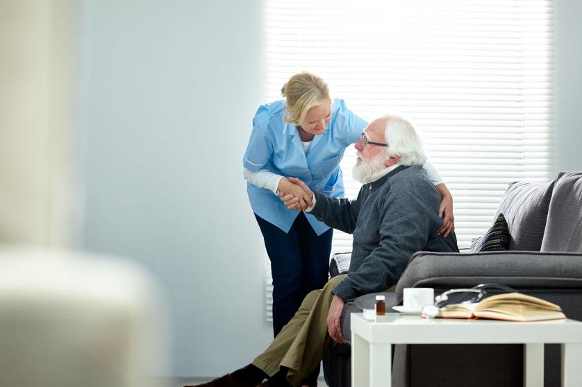 Female caregiver helping senior man get up from couch in living room