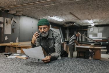 Szakállas férfi tulajdonos tervrajzzal beszélget az okostelefonon keresztül, miközben a munkapadon támaszkodik
