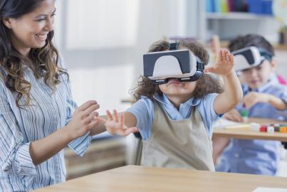 Cheerful mid adult Asian teacher helps young female student use virtual reality glasses in class