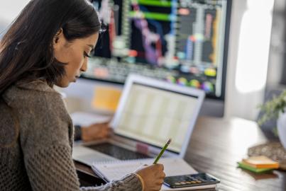 Middle Eastern woman tracking and trading stocks using laptop and desktop computer. 