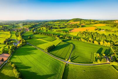 Aerial panorama over picturesque river valley meandering between rolling hills of patchwork pasture, agricultural crops, rural homes and green summer landscape