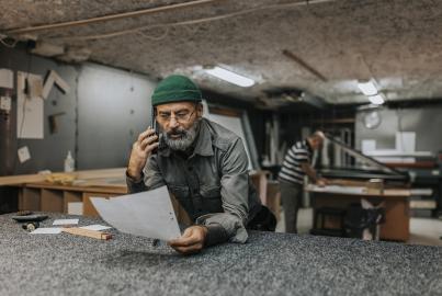 Bärtiger männlicher Besitzer mit Blaupause, der über sein Smartphone spricht, während er sich auf eine Werkbank stützt
