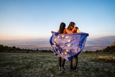 Two young pretty multi-ethnic women holding European Union flag on meadow