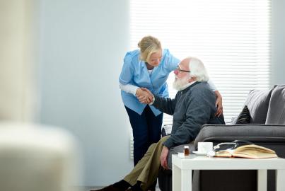 Female caregiver helping senior man get up from couch in living room
