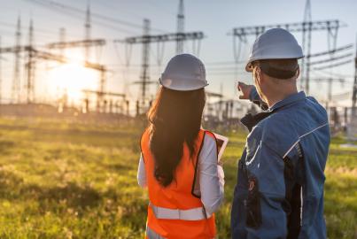 Um operário aponta para os postes de eletricidade, com o pôr do sol ao fundo, ao lado dele está uma engenheira