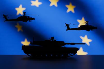 Una gran bandera de la UE al fondo, con equipo militar moderno en primer plano, visto en silueta