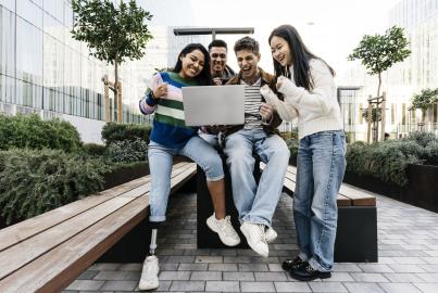 Multiracial group of young students outside on campus or university looking at laptop and sitting on top of a table celebrating good news.